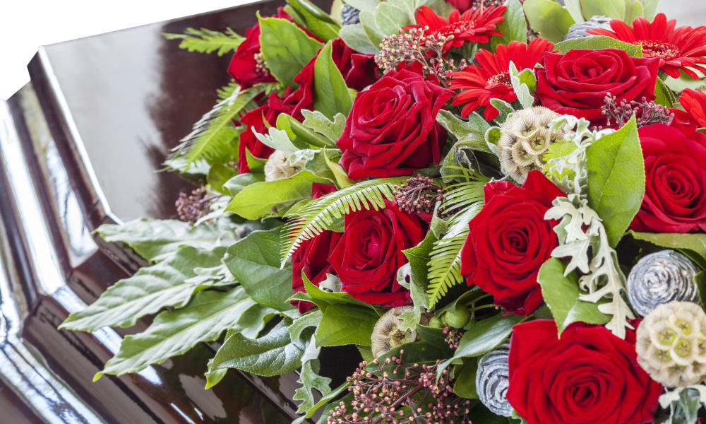 A close up image of a casket with a beautiful arrangement of Red Roses and greens. very rare white and purple flowers too