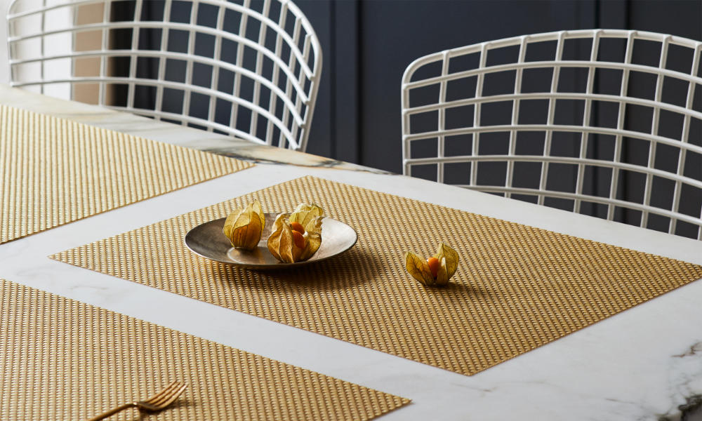 A dining table setting with very stylish and modern golden placemats, a small desert plate and a desert fork on one of the placemats, and two contemporary dining chairs on the back