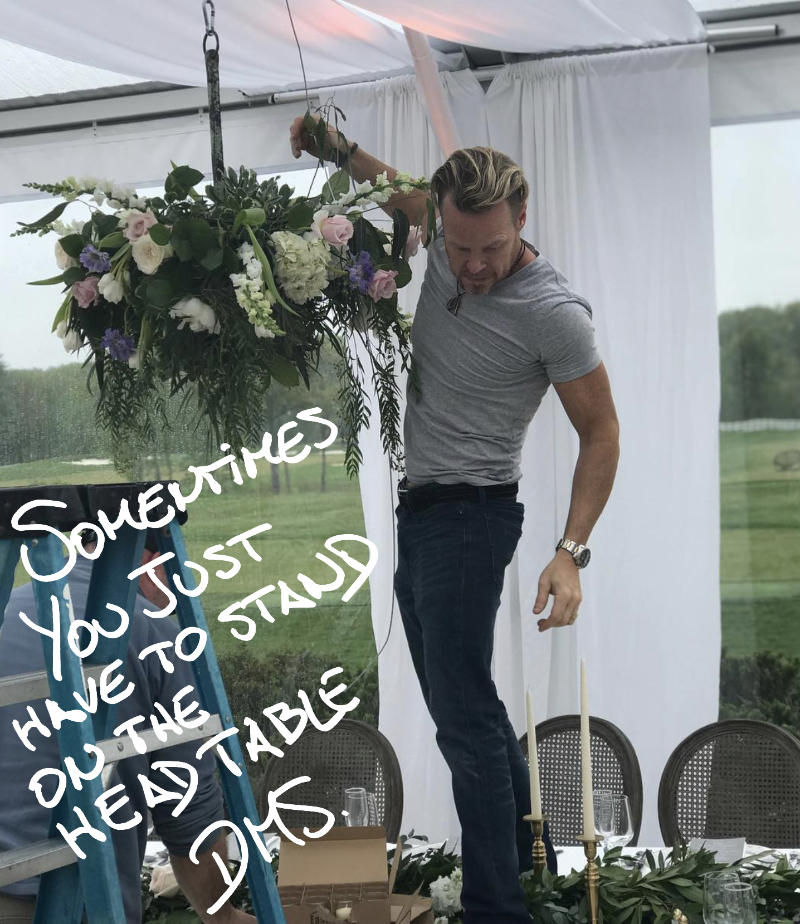 A handsome guy standing on a head table of a wedding hanging a stunning wedding flowers from the roof of a white tent