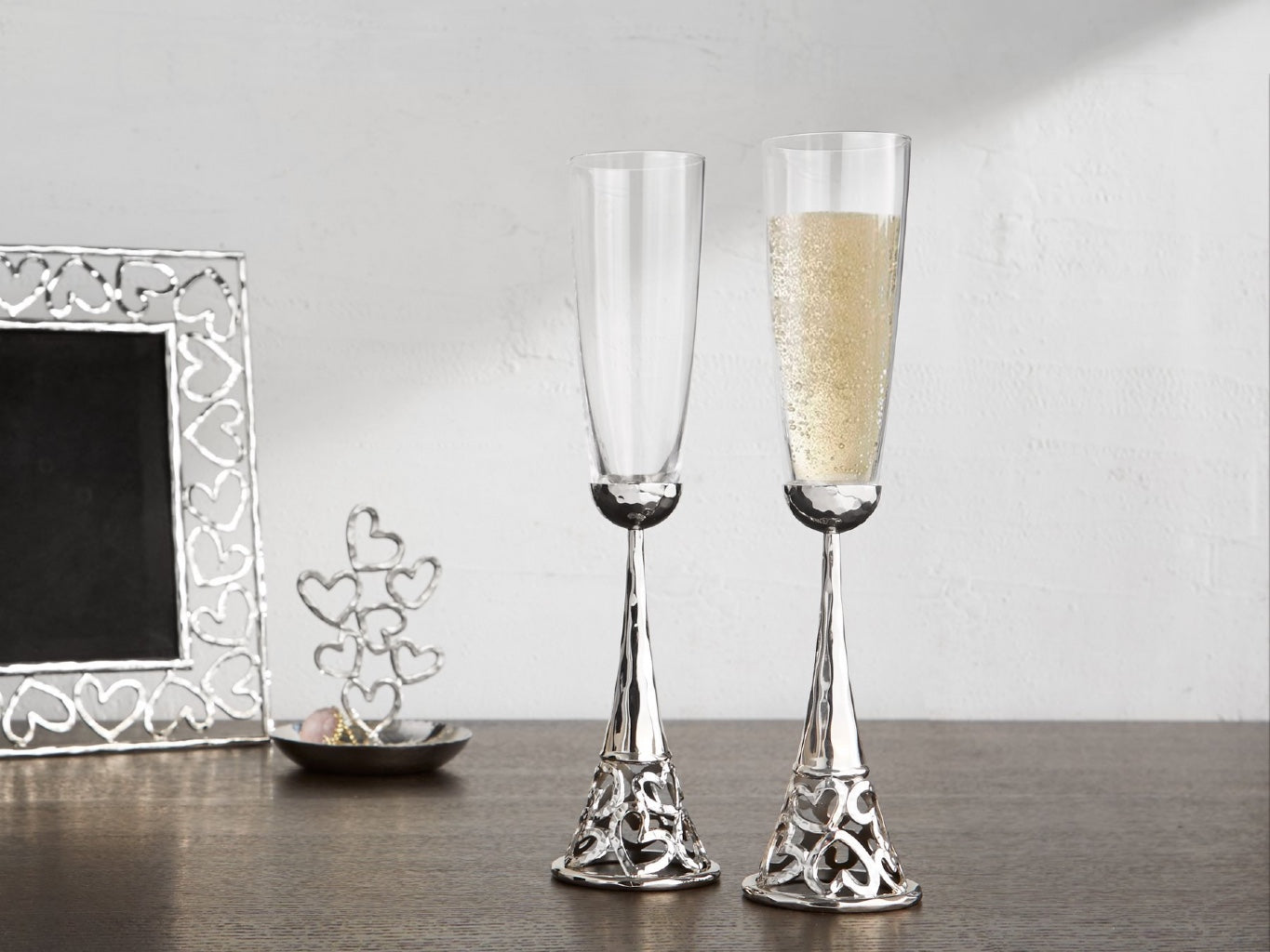 Two toasting flutes with a silver vase, designed with heart shape on the bottom, a picture frame with the same hearts shape and silver finish in the background, and a small sculpture with stacking little hearts.