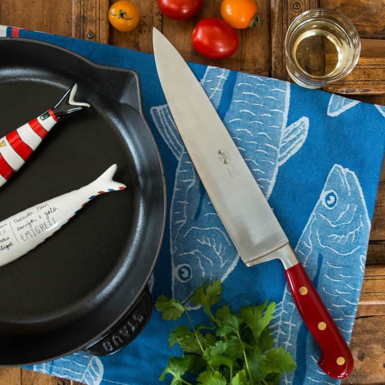 A Red Lucite Kitchen Knife next to a cast-iron skillet on a cloth placemat with a fish pattern. Some tomatoes are on the table.