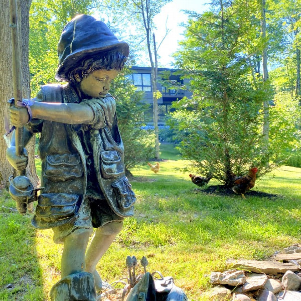 A fisher boy bronze statue in a garden, a modern house in the background, and some chickens are playing around the yard. 