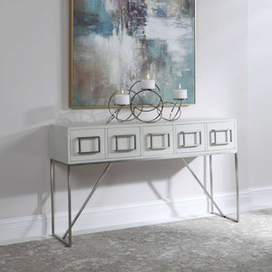 A white sideboard table, with doors and square silver handles, silver finish legs, and a set of candles on top of it.