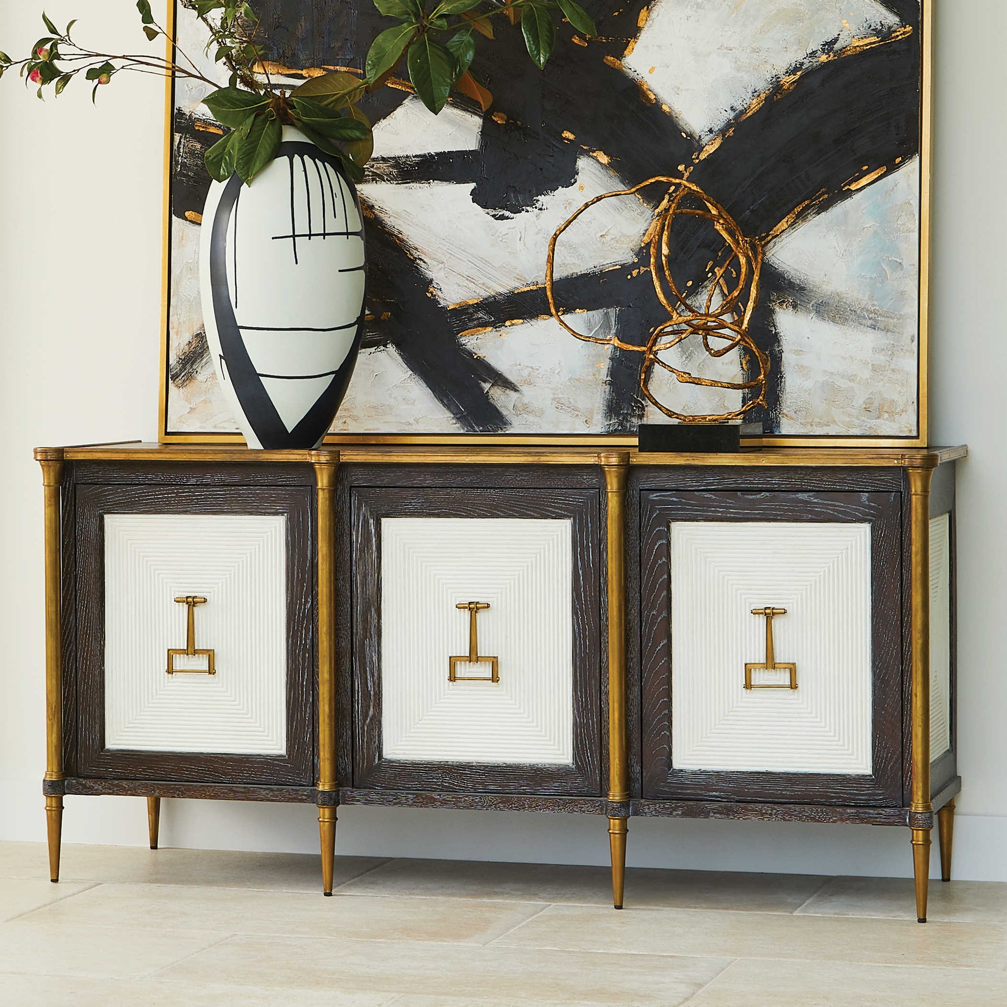 Beautifully detailed cabinet, with a gold finish and classical elements. A modern sculpture and a black and blue bottle on it. And an abstract artwork giving a modern look to the scene.