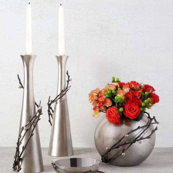Two tall and very modern candleholders in a silver plated material, nest to them a bowl filled with red roses