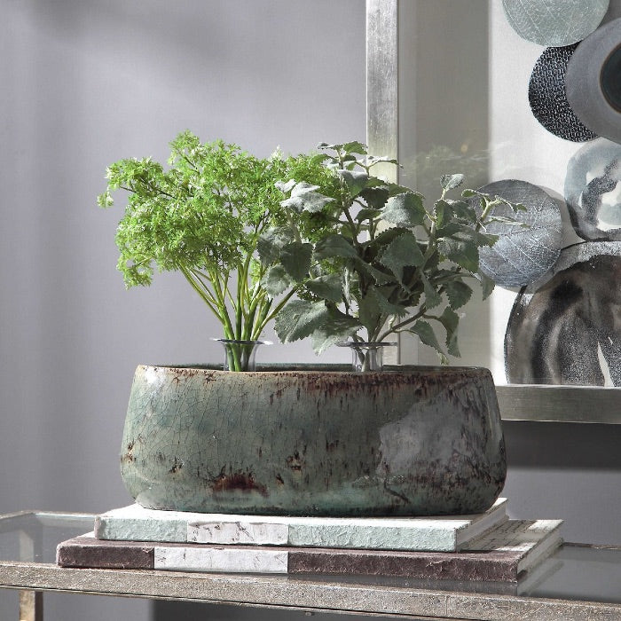 A Ceramic bowl finished in an aqua blue crackled glaze with chocolate brown accents, on top of a console table, and two large books. It contains two green plants, and there is a shadow box hanging on the blue wall in the background.