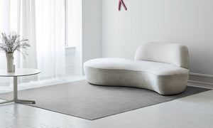 A minimalistic and modern living room, with only three pieces: a modern cozy white couch, a round side table with some flowers, and a beautiful woven floor mat. The place is stunning and neat