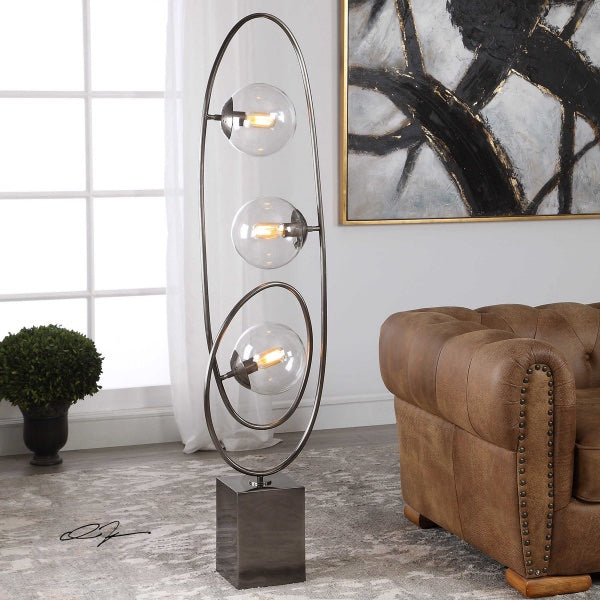 A modern floor lamp design features an abstract steel construction finished in a lightly antiqued plated brushed nickel, surrounding three clear round glass globes, next to a leather couch, and on the background an abstract artwork.