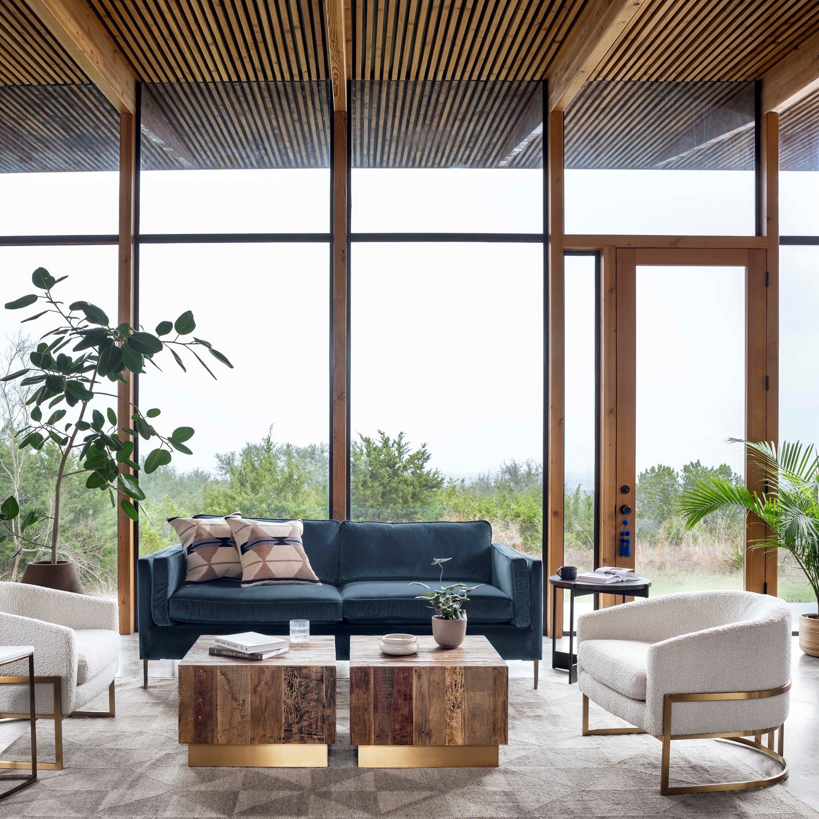 A stylish living room with floor to ceiling windows with a garden view, in the room two white accent chairs with golden details, a blue sofa and two cube tables with golden details, some green plants bring the outside garden in.