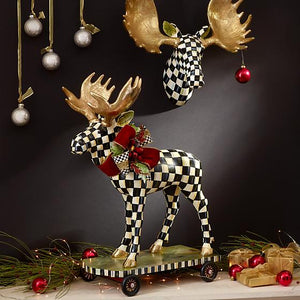 A very colorful holiday decor scene shows a courtly check moose with golden antlers on a rolling skate, on the black wall a moose head with impressive and very big golden antlers.