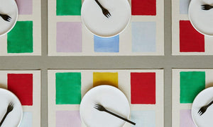 A picture from above of a dinning table with six placemats with colorful squares shapes. and six empty dessert plates with black modern forks