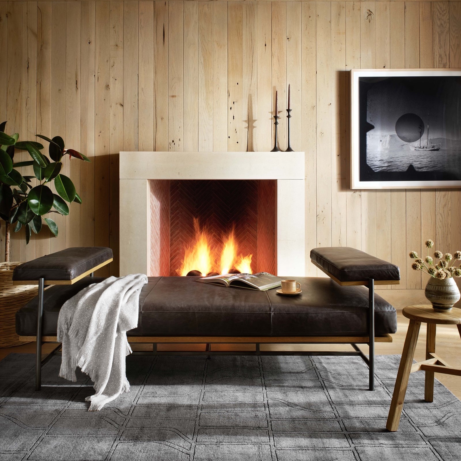A cozy room with a fireplace and a brown leather couch, with a white blanket on top of it. An accent stool has a cocktail with an arrangement.