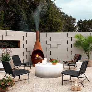 A Modern patio with Scandinavian style grey walls, an iron cast fireplace, four outdoor chairs, a round coffee table, and an end table, the floor is cover with some kind of stones, and there are some green plants around the patio.