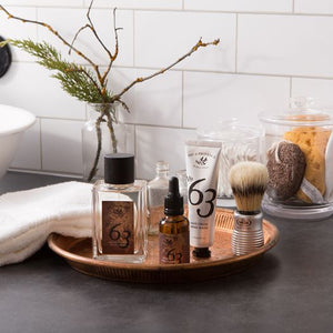 A copper color vanity tray with shaving items on it, and a white face towel and a vase with a plant on the same countertop.