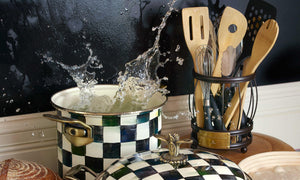 A large pan and a pot on a stove, water splashing out from the large pot, and a set of Mackenzie Childs utensils next to the stove