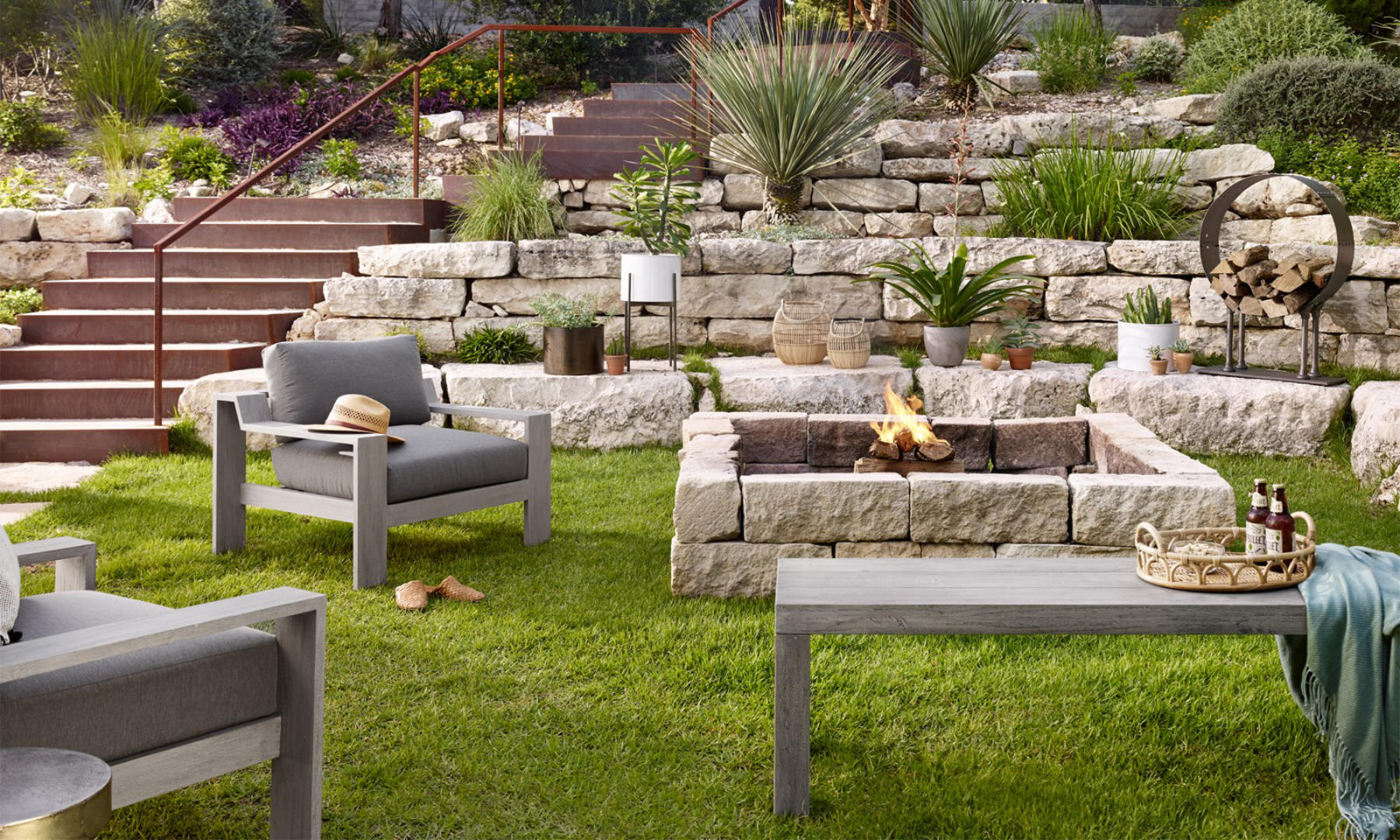 A stunning patio set up, with grey furniture, a fireplace, lots of pots and plants, and a stone wall on the back.