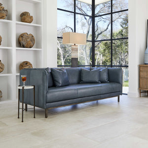 Featuring a blue leather sofa, a drink table with a wine glass on it. and behind an accent lamp and a ceiling to floor window