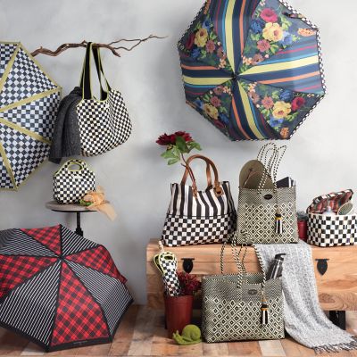 Different bags and totes around a foyer wood bench and on the floor and hanging from a decorative tree branch, also there are three umbrellas in the scene. All the items have colorful patterns with flowers, stripes, and dots.