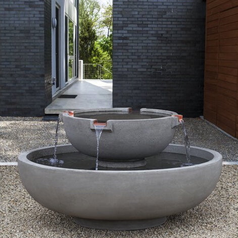 A cast stone round fountain with a modern and minimalistic style, placed over the gravel patio floor of a modern house, with a black bricks wall on the background.