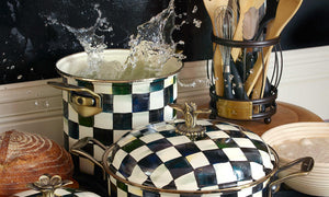 A couple of  Mackenzie Childs pots on a stove, one of the pots have water splashing out of it. all the cookware have the distinct Courtly check Pattern