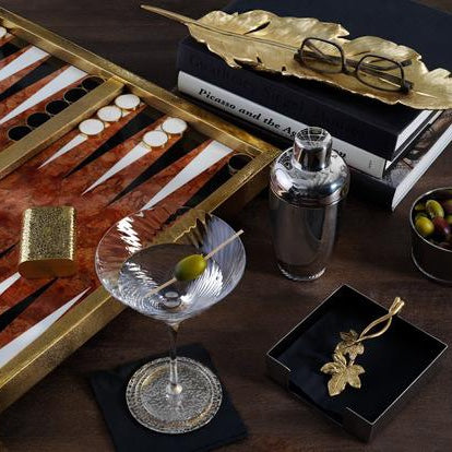 A backgammon board with some pieces on place, which tells you the game has started, there is also a martini glass with a cocktail served in it, a small bowl with olives, and some napkins on the table.