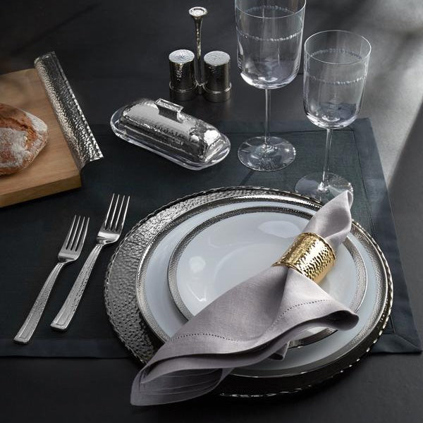 A dining table set up, with a black fabric placemat, two glasses, white plates, a fashion golden napkin ring, and a grey cloth napkin.
