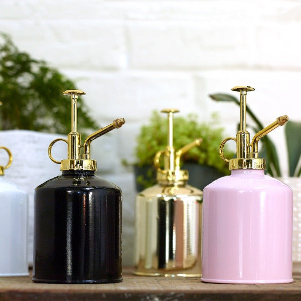 Set of fashion watering cans, gloss metal finish in pink and black on the front and a golden and a white on the back, all of them with golden finish ion the tip. Some planter and plants in the background.
