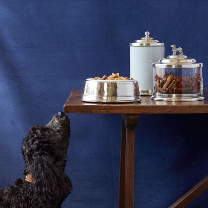 A table with a pet bowl and two pet food containers and a dog is trying to reach the food that is on the table.