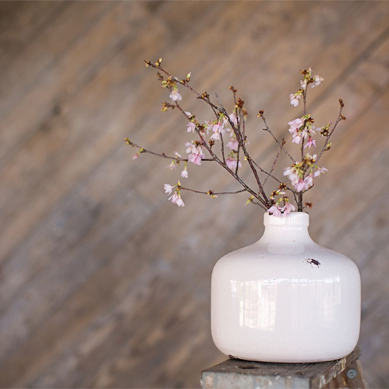 A white ceramic bottle with some brown details, on top of an old fashion wood ladder. It contains an arrangement of little pink flowers. The background is a blurry brown wall