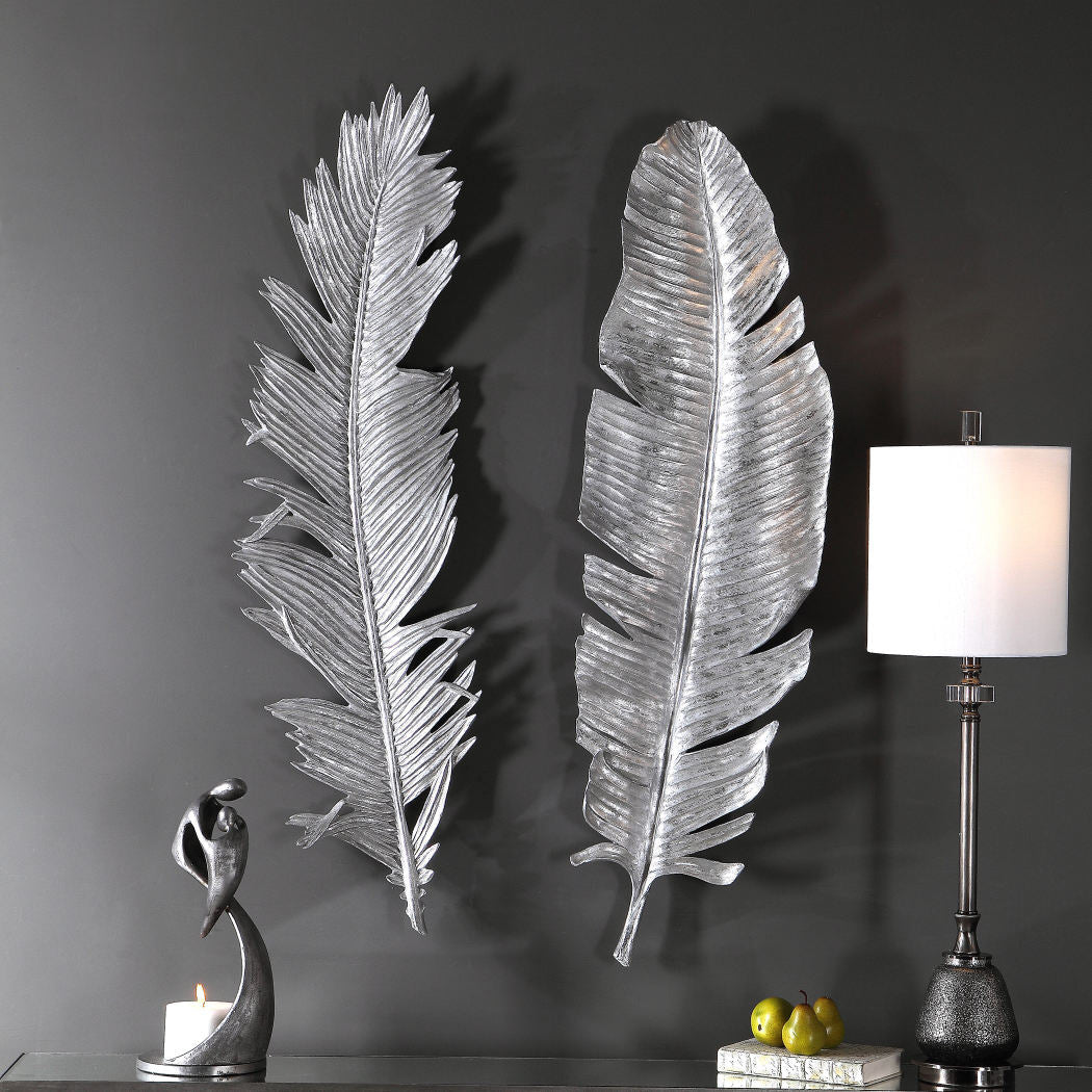 A grey wall featuring two beautiful and stylish metal leaves, these pieces of wall art are big and shiny, a small statue and a lamp on a table complete the gorgeous scene 