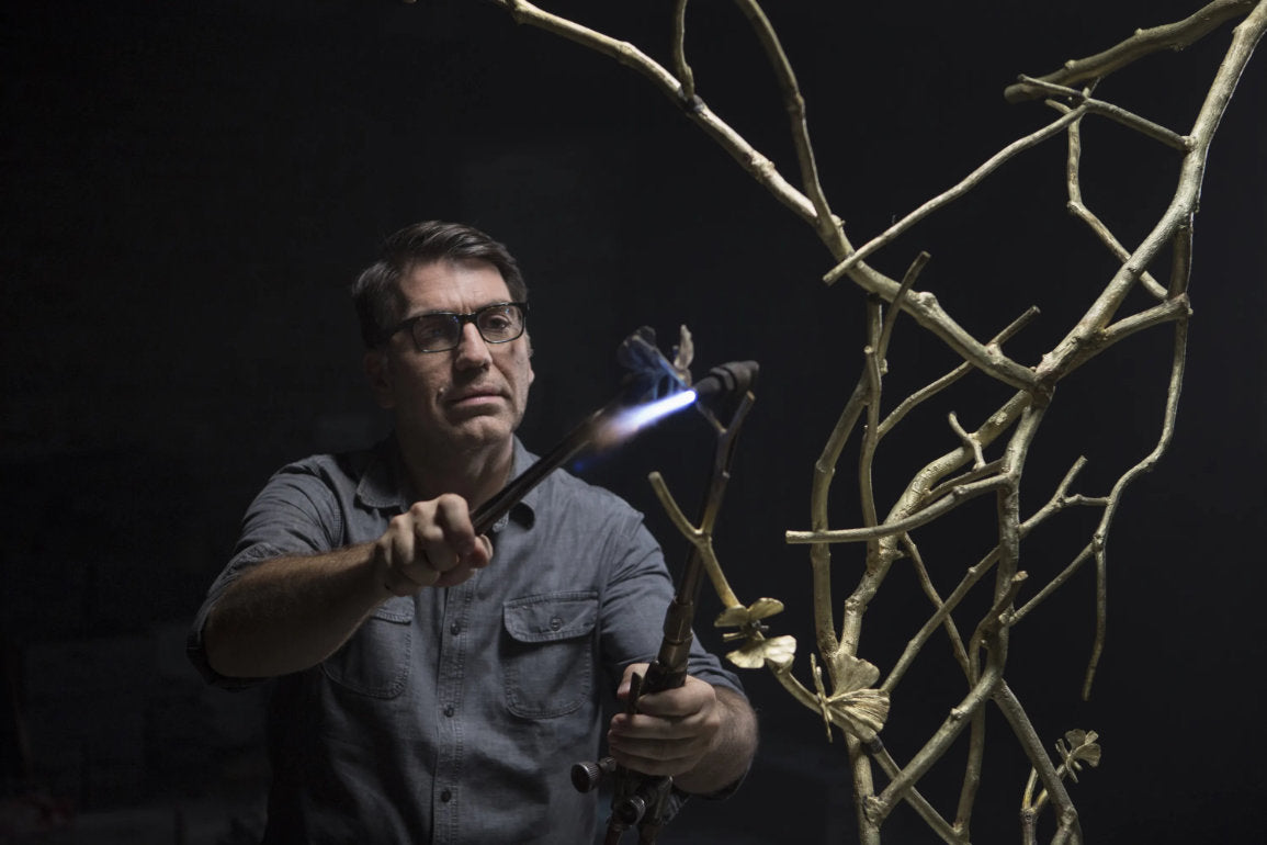 Michael Aram, the artist, is welding some pieces of metal to creat an sculpture representing branches of a tree with butterflies on it