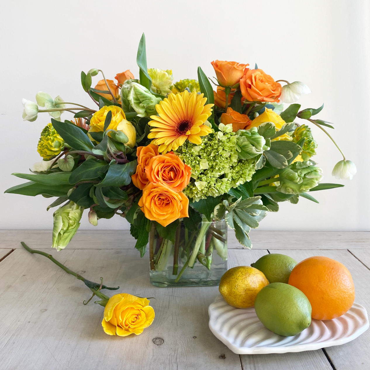 A beautiful citrus toned flower arrangement. A mix of lemon, limes, bright oranges, and stunning yellows. The arrangement features green Hellebore, green and white Parrot Tulips, orange crush Spray Roses, Kiwi Hydrangea, and large two-toned Gerbera Daisys