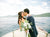 bride and groom in a boat around the lake in abeautiful sunny day, holding a geaourgous white bouquet