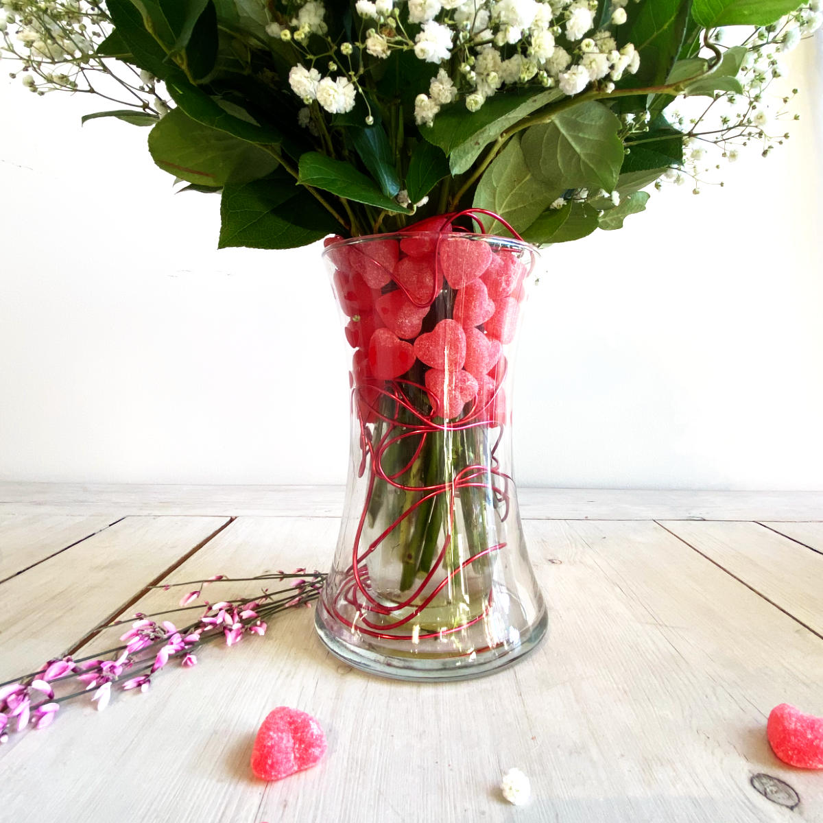 A dozen lush red roses and bb in a double vase with Red Hot Cinnamon Candies inside the vase and a red wire. the candies have a heart shape. also some candies on the table
