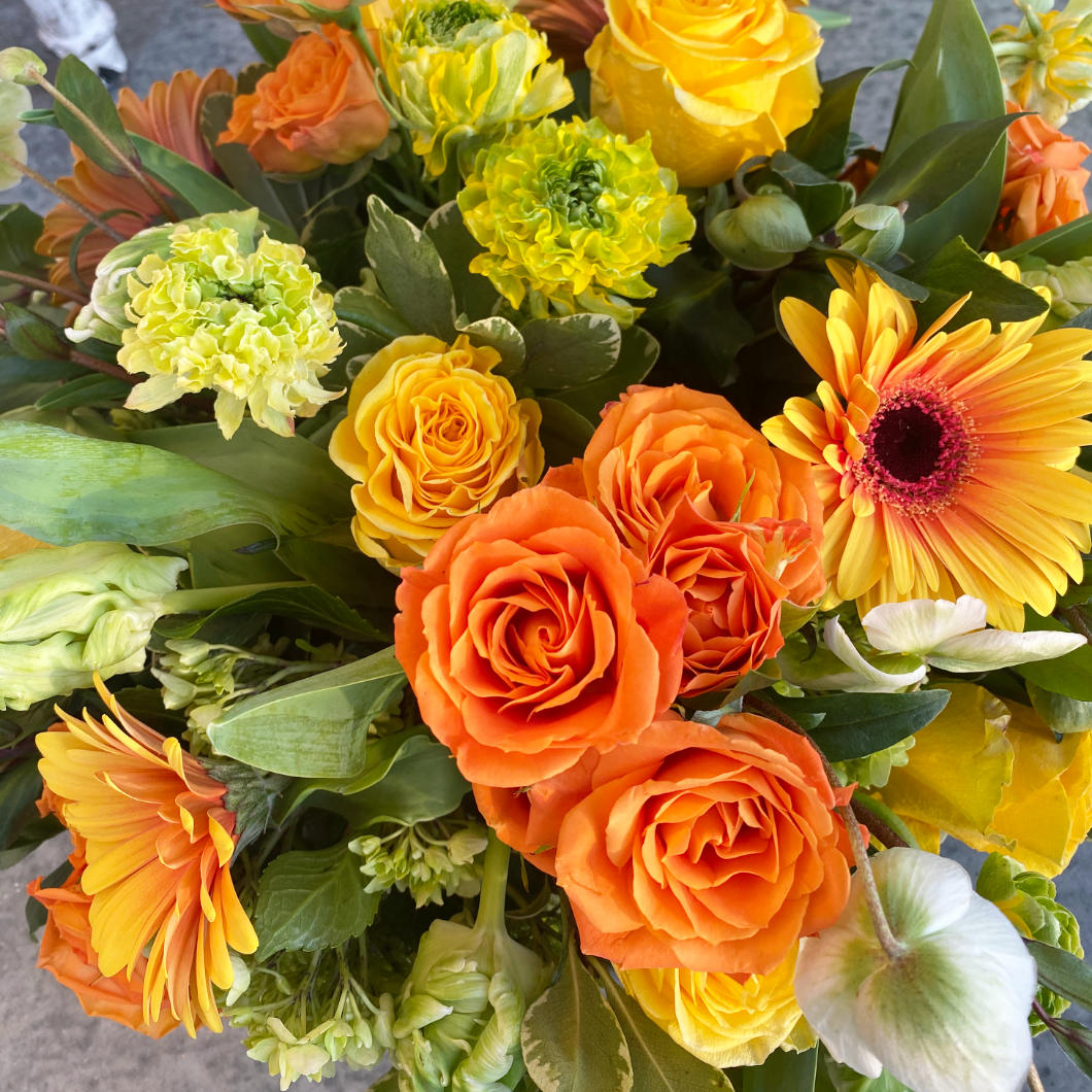 A beautiful citrus toned flower arrangement. A mix of lemon, limes, bright oranges, and stunning yellows. The arrangement features green Hellebore, green and white Parrot Tulips, orange crush Spray Roses, Kiwi Hydrangea, and large two-toned Gerbera Daisys. In a clear glass cylinder. next to it, a plate with lemons and limes, an