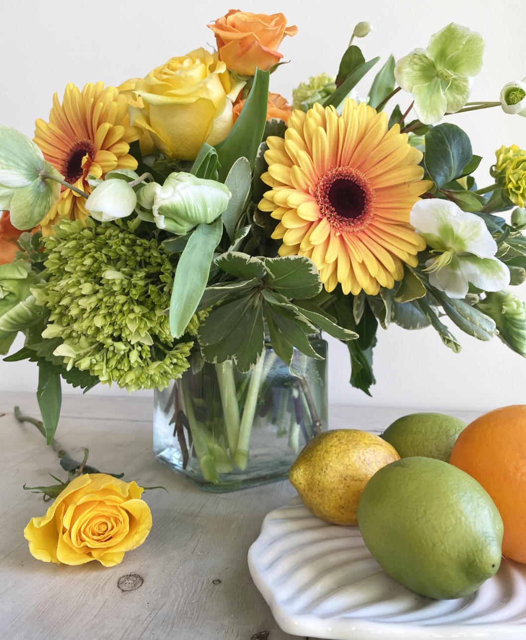 A beautiful citrus toned flower arrangement. A mix of lemon, limes, bright oranges, and stunning yellows. The arrangement features green Hellebore, green and white Parrot Tulips, orange crush Spray Roses, Kiwi Hydrangea, and large two-toned Gerbera Daisys. In a clear glass cylinder. next to it, a plate with lemons and limes, and there is a yellow rose over the table