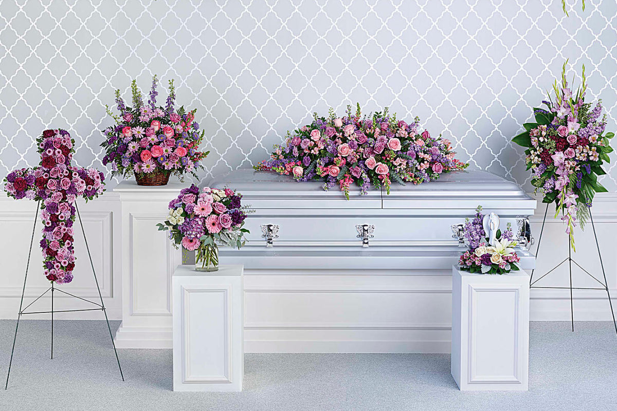 A collection of funeral flower arrangements, featuring six pieces with pink, red and purple roses. all the flowers around a grey casket in a grey room of a funeral home.