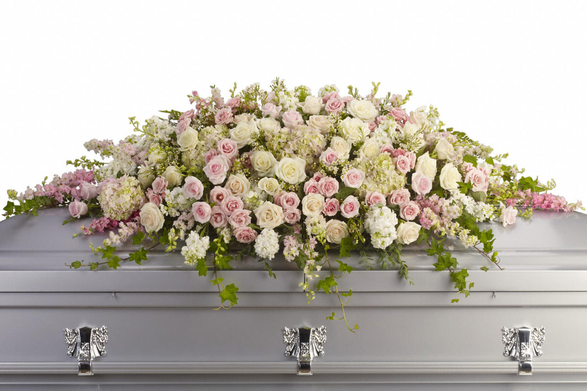 A casket spray made with whit and light pink roses on top of a grey casket