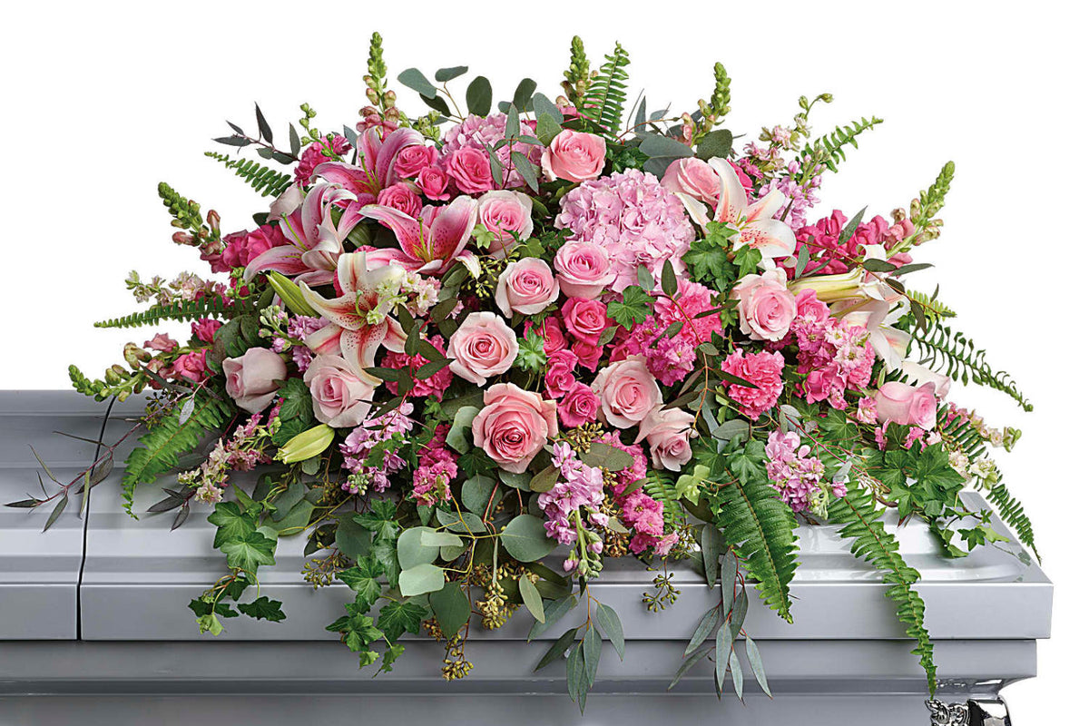 A dramatic and beautiful casket spray features pink hydrangea, pink roses, pink spray roses, pink stargazer lilies, pink carnations, pink larkspur, pink snapdragons, pink stock, green ivy, sword fern, silver dollar eucalyptus, seeded eucalyptus, and lemon leaf. The funeral flowers stand on top of a grey closed casket