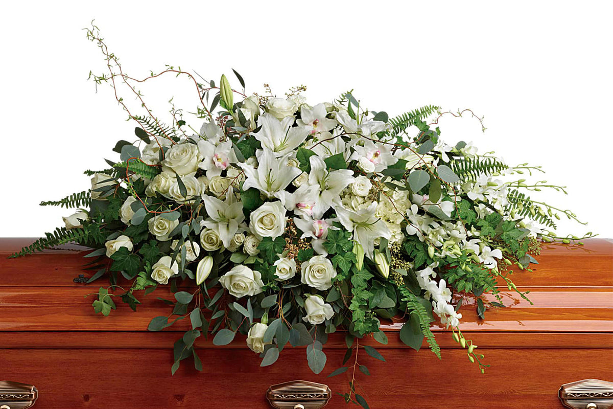 Funeral flowers, specifically a casket spray with green almost white  roses and white lilies, and a lot of greens, over a brown casket