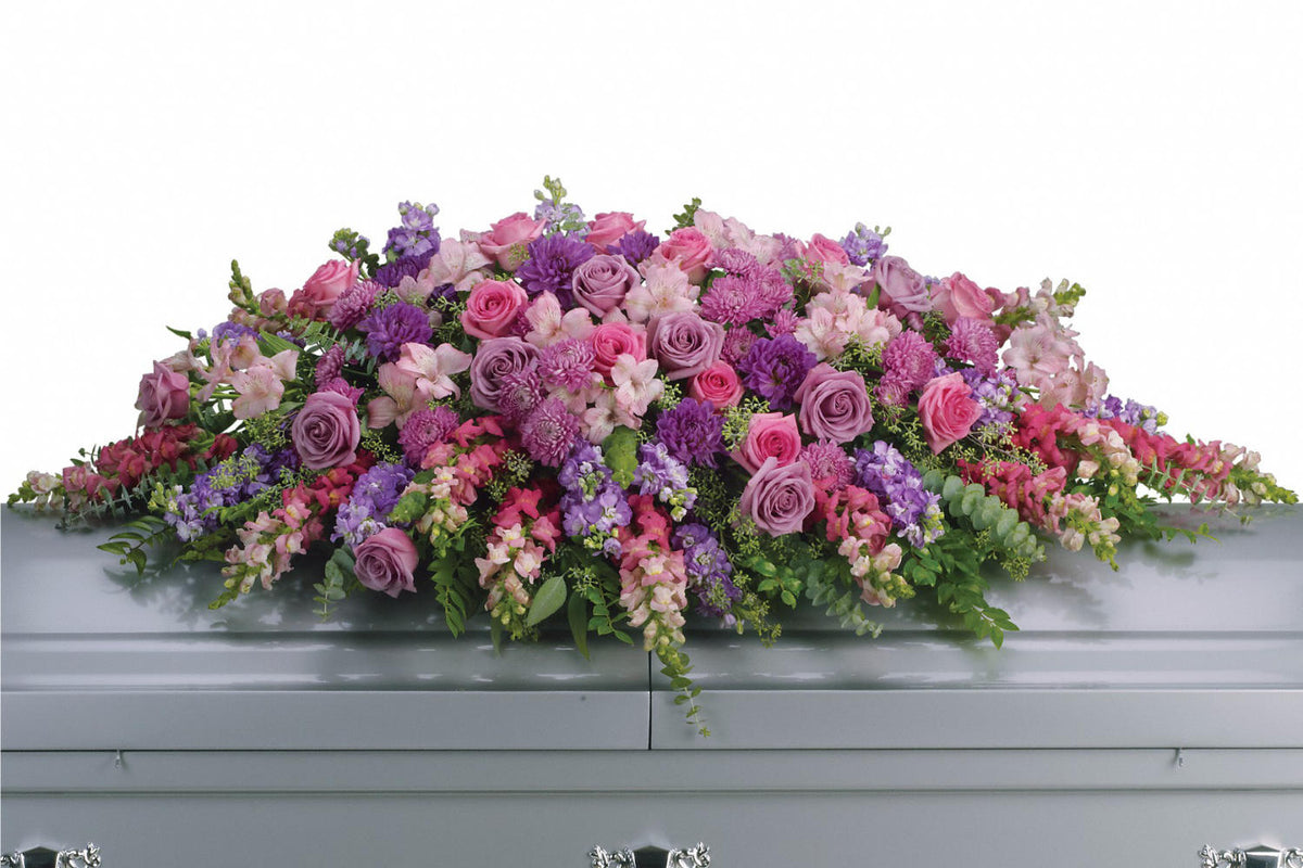 Funeral flowers arrangement with lavender and pink roses, snapdragons, alstroemeria, chrysanthemums, fern, eucalyptus and more, all of it create this tribute that is overflowing with grace and love over a metallic grey casket