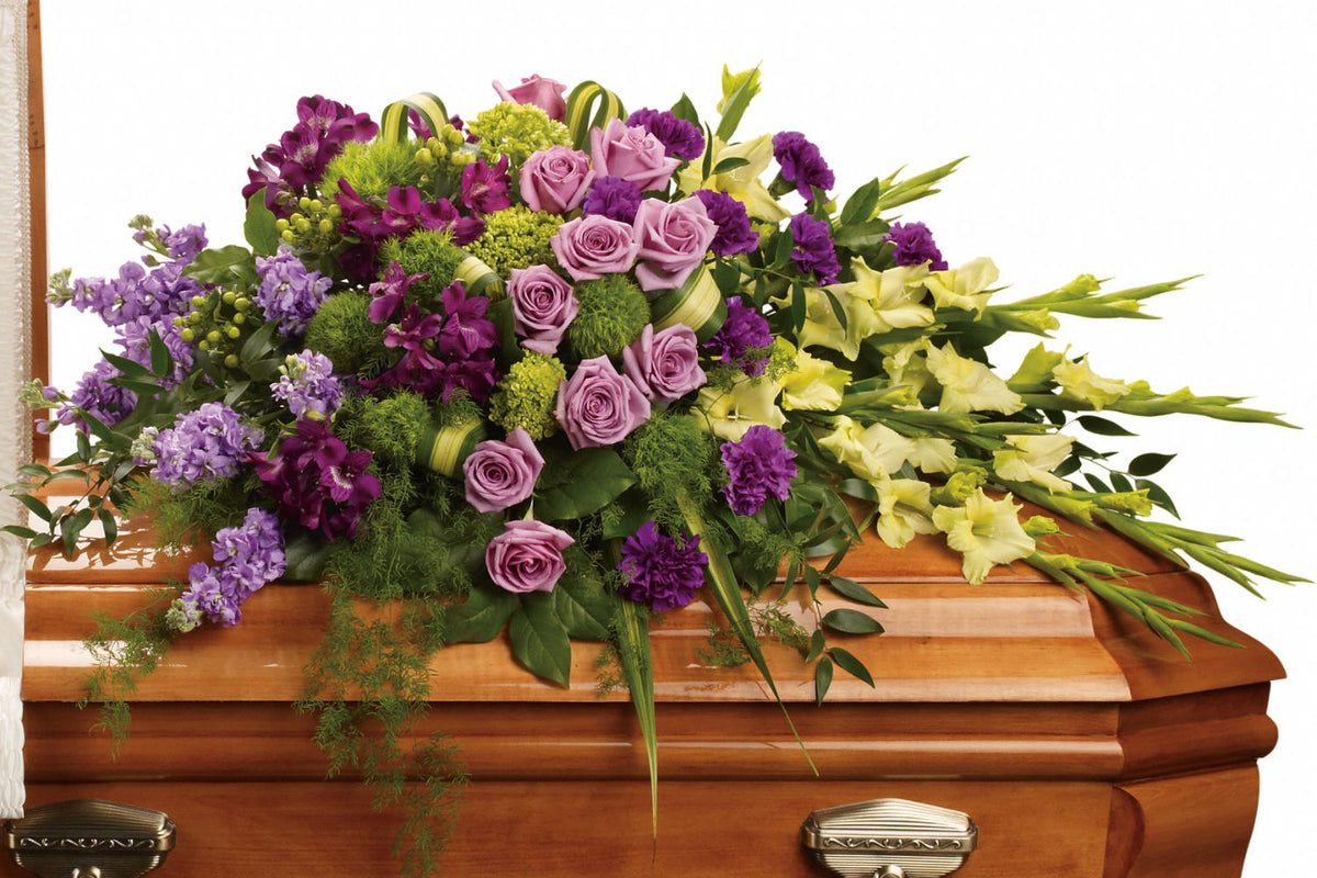 Funeral Flower arrangement. The lush arrangement includes green miniature hydrangea, lavender roses, purple alstroemeria, green gladioli, green trick dianthus, purple carnations, lavender stock and green hypericum, accented with assorted greenery. the casket spray is on a brown casket