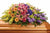 Colorful funeral flowers in a casket spray, it has pink lilies, pink roses, peach roses, yellow flowers, purple hydrangea and green lilies, over a brown casket