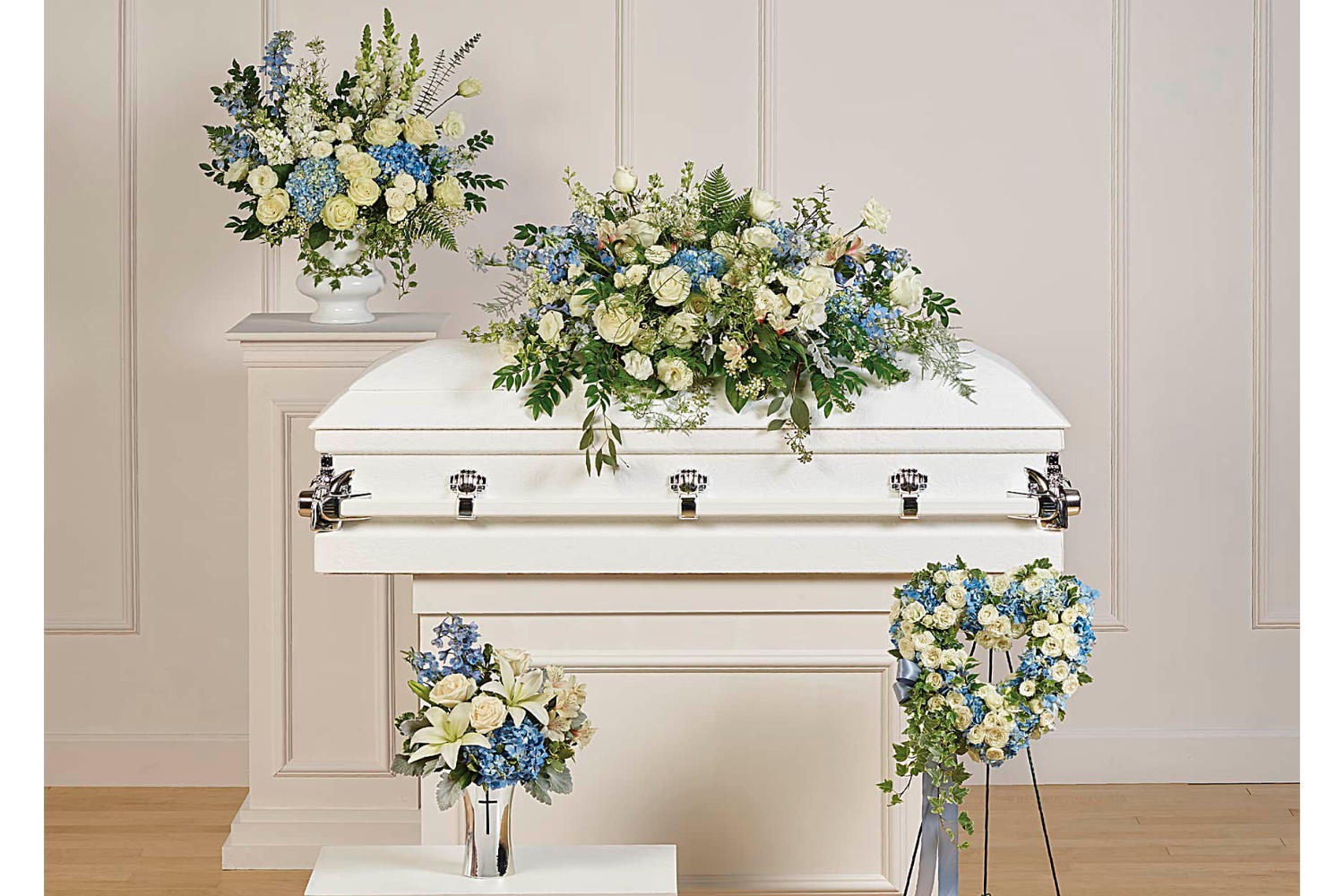 A collection of four hand-made sympathy pieces in white roses and blue hydrangea. all the funeral flowers arrangements are set up in a funeral home around a grey casket.