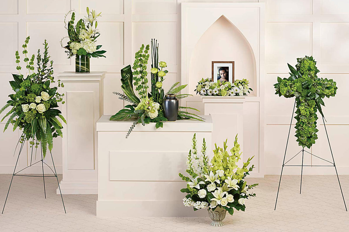 Calming white blooms and unique, sculptural greens give this collection of six hand-made sympathy pieces a tranquil, zen-like feel. all the funeral flowers arrangements are set up in a funeral home around a grey casket.