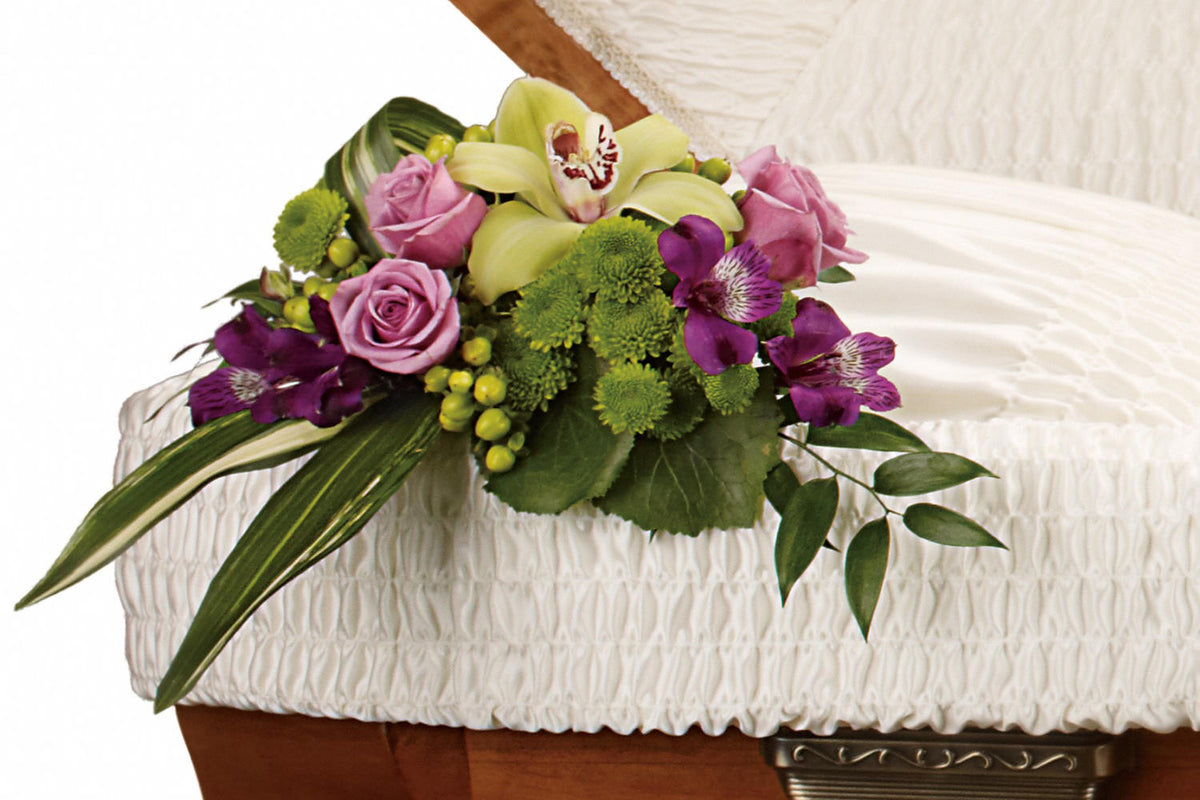 A Funeral flower  arrangement placed on the corner of a casket. have orchids, pink rises and beautiful greens