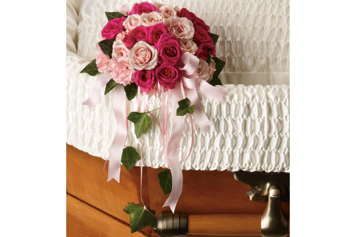 A delicate bouquet includes hot pink spray roses and light pink miniature carnations tied with pink satin ribbon, accented with green ivy. this funeral flower arrangement is hanging on the corner of an opened brown casket