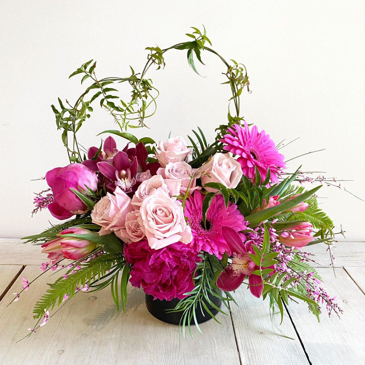 A beautiful flower arrangement in a gloss black vase, featuiring pink roses, orchids and gerbera daisy flowers