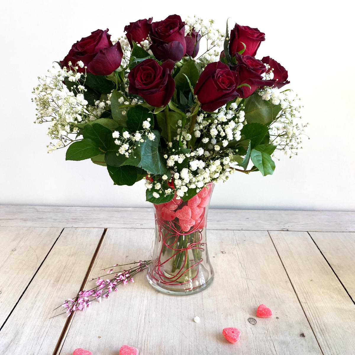 A dozen lush red roses and bb in a double vase with Red Hot Cinnamon Candies inside the vase and a red wire. the candies have a heart shape. also some candies on the table